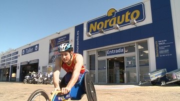 Un deportista con discapacidad física posa, sobre su silla de ruedas deportiva, junto a una sede de Norauto