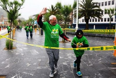 Una persona mayor cruza la meta bajo la lluvia junto a su acompañante, un niño provisto de antifaz que le impedía la visión