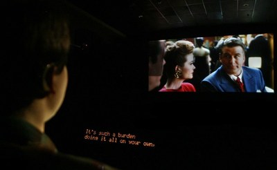 Una persona sorda (en primer plano) contempla una película en una sala de cine siguiendo los diálogos gracias al subtitulado