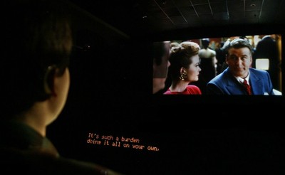 Una persona sorda (en primer plano) contempla una película en una sala de cine siguiendo los diálogos gracias al subtitulado