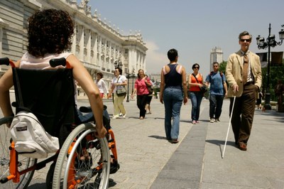 Una mujer en silla de ruedas se cruza con un hombre ciego que camina con su bastón en la explanada de acceso al Palacio Real en Madrid