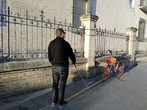 Una persona ciega camina por una acera en la que hay aparcada/abandonada una bici de alquiler compartido 