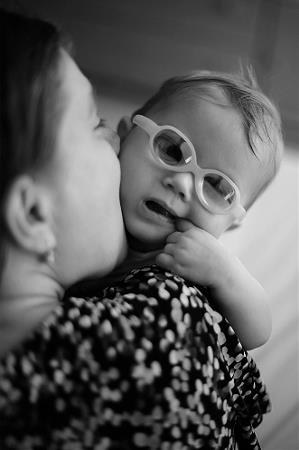 Madre con discapacidad visual besa en la mejilla con la dulzura, en un gesto de amor incondicional, a su bebé con gafas menor de un año. Foto de Ariel  B. Pascuali