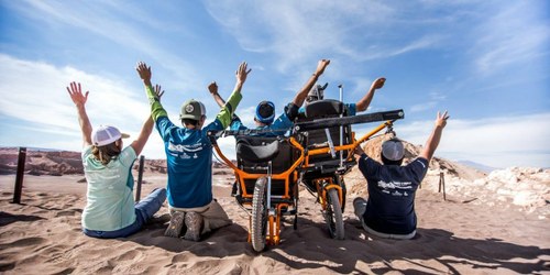 Un grupo de jóvenes con discapacidad disfrutan sobre la arena de la playa, de espaldas, junto a sus sillas de ruedas