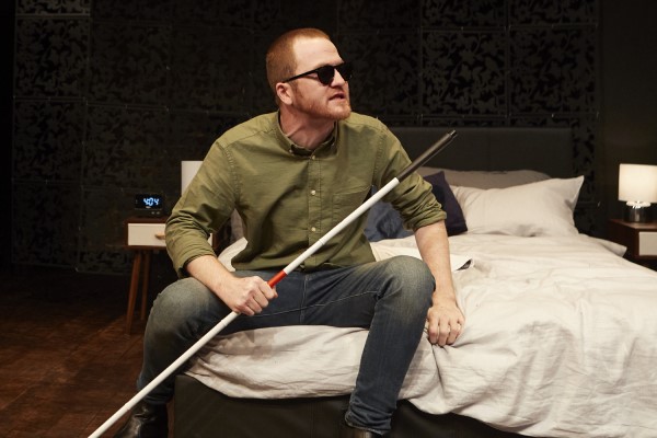 Sam Brewer con un bastón blanco en la mano, sentado en la cama, durante su representación
