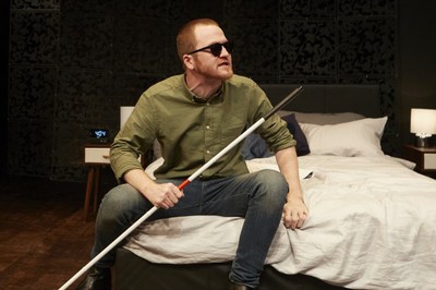 Sam Brewer con un bastón blanco en la mano, sentado en la cama, durante su representación