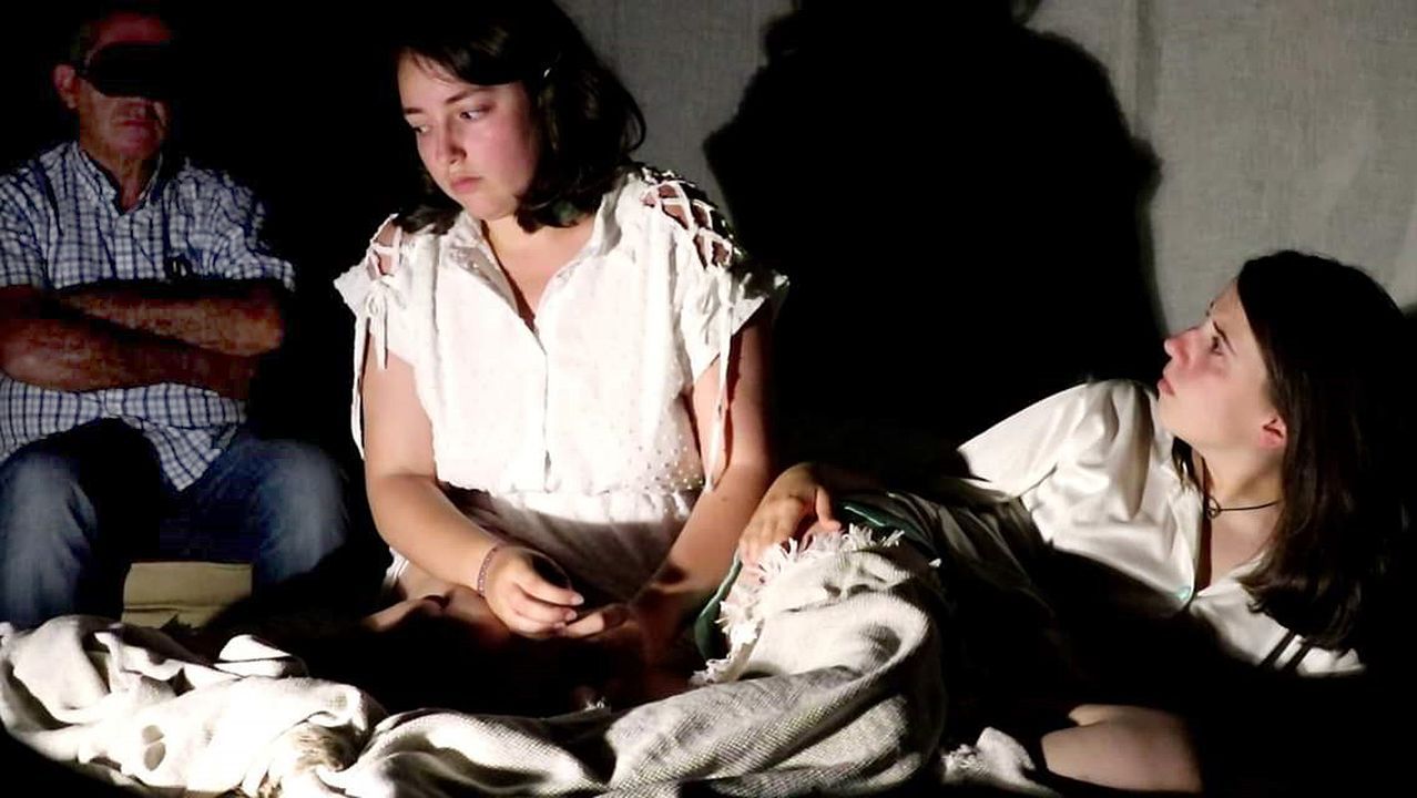 Momento del montaje "Coa mirada nas súas mans" con actrices en primer plano y espectador al fondo con antifaz