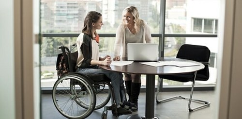 Mujer en silla de ruedas en su oficina de trabajo junto a una compañera sin discapacidad