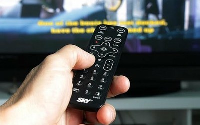 Pantalla de televisor al fondo de la imagen, con una mano que sujeta un mando a distancia en primer plano