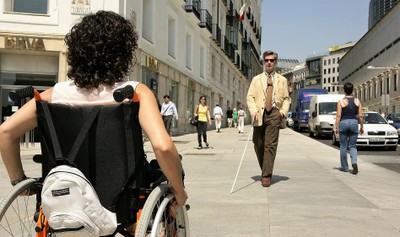 Una mujer en silla de ruedas y un hombre ciego, con su bastón, se cruzan en una acera de una ciudad española