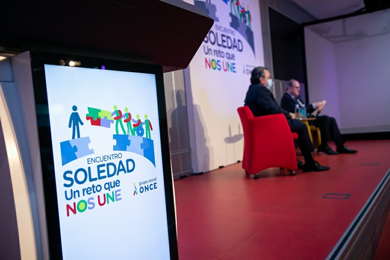Primer plano del cartel de la jornada con Carballeda y Gabilondo al fondo en el acto de inauguración