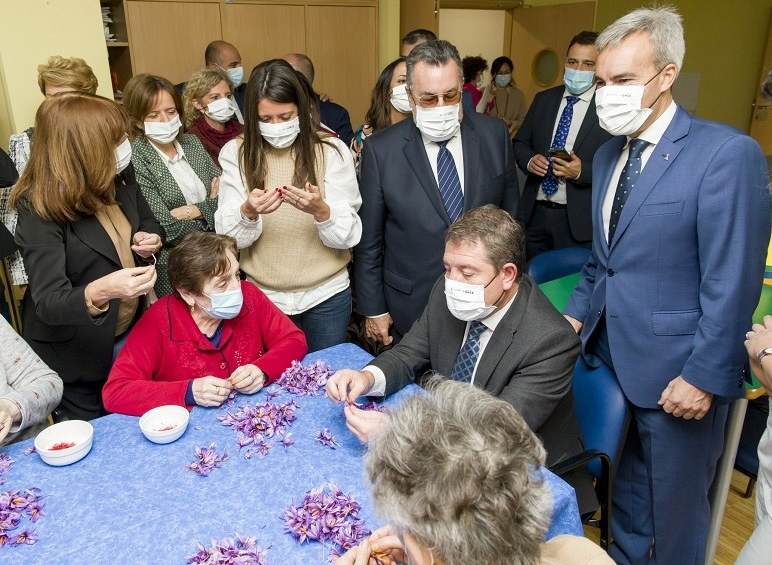 García-Page sentando con las residentes sacando hilos de las flores del azafrán, copn Miguel Carballeda, de pie, detrás