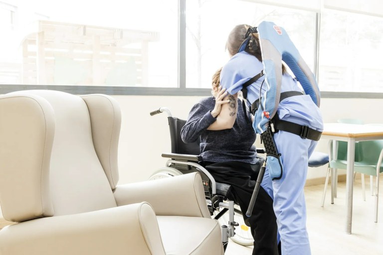 Trabajadora de Ilunion con un exoesqueleto levanta de su silla a una persona mayor
