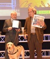 El escritor Jaume Cabré, homenajeado en la gala, recibe, de manos del delegado territorial de ONCE en Cataluña, sendos ejemplares en versiones braille y sonora de una de sus obras