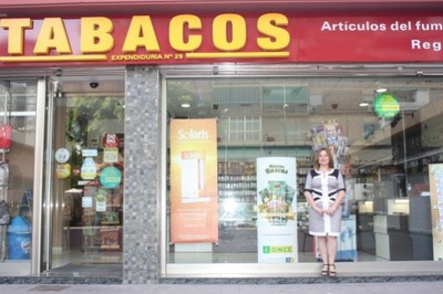 María Jesús, ante la amplia fachada de su estanco, lcuiendo varios carteles promocionales de nuestros productos