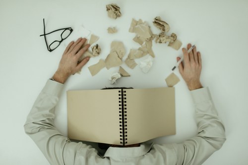 Hombre agotado bajo cuaderno, folios arrugados y útiles de escritura